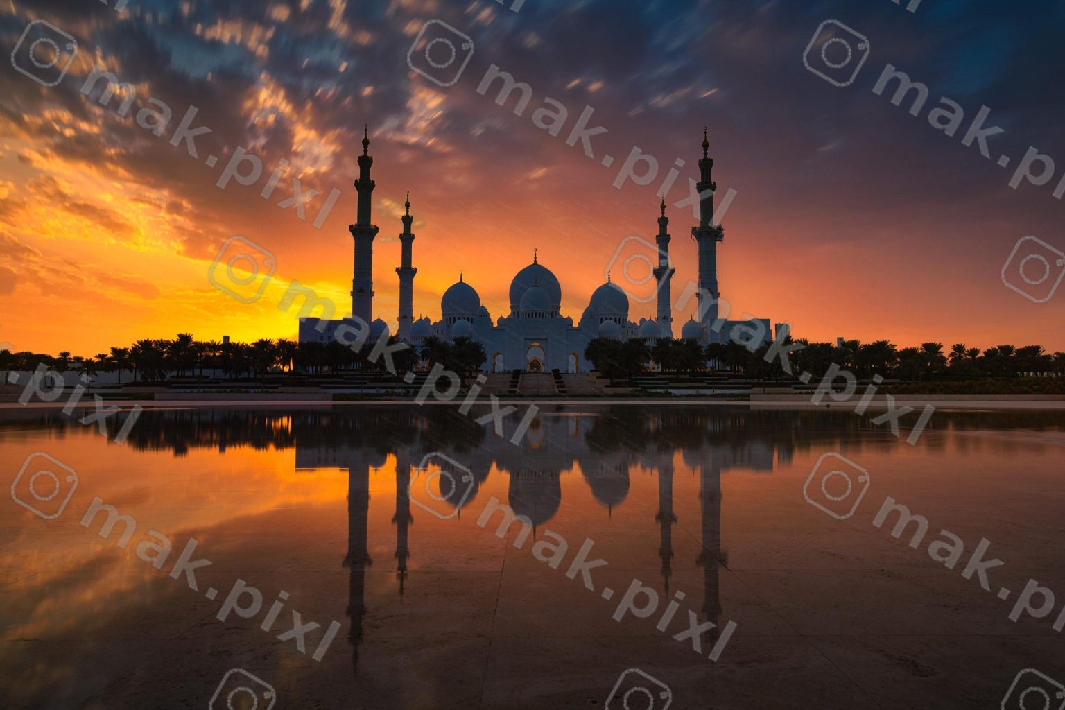 A grand mosque with multiple domes and minarets is silhouetted against a vibrant, colorful sunset sky, with its reflection visible in a still body of water in the foreground.